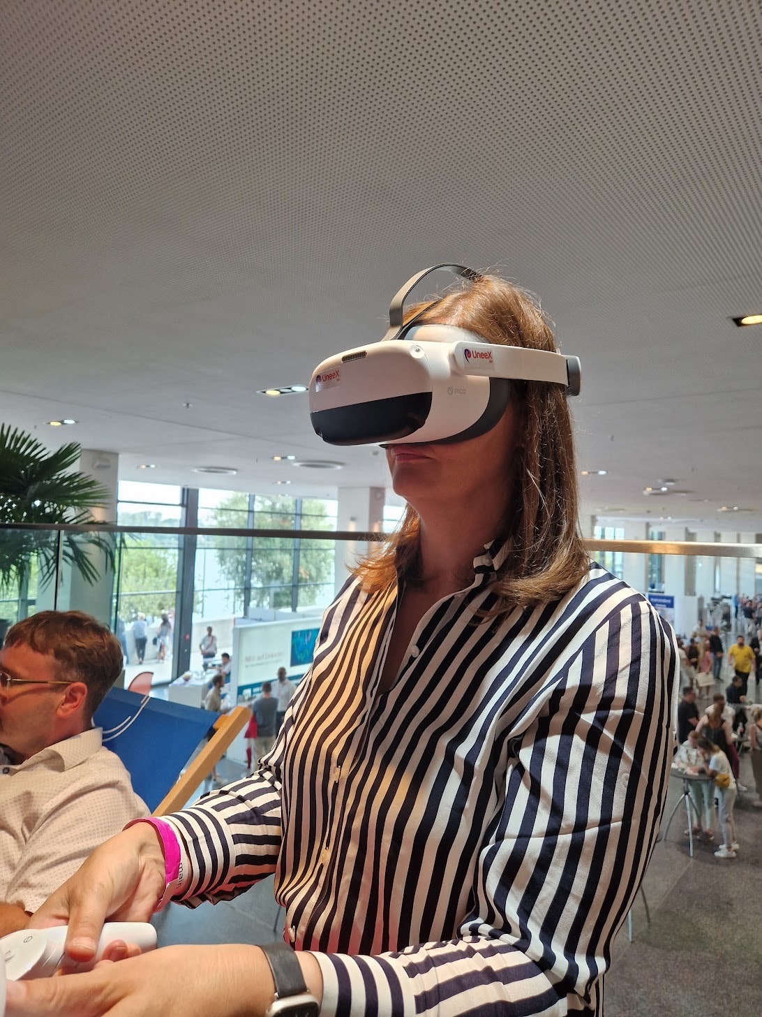 Frau in gestreifter Bluse Teste eine VR-Brille auf einer Messe. Im Hintergrund weitere Personen die an verschiedenen Tischen stehen.